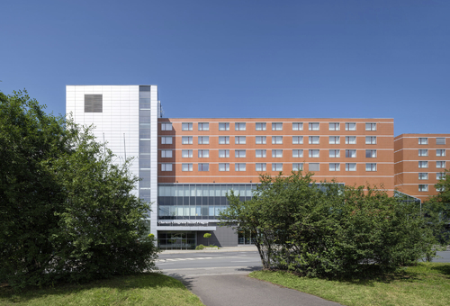 Hotelgebäude mit modernem Design, umgeben von Bäumen und blauem Himmel.